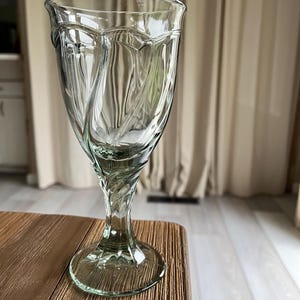 May include: A clear, elegant water goblet with a decorative, swirling design. The glass has a wide, oval-shaped top and a detailed stem. The goblet is made of clear glass and sits on a wooden table.