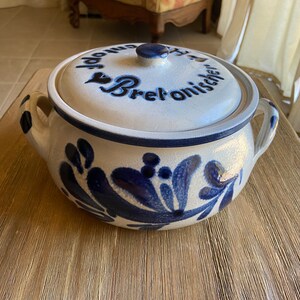 May include: A white ceramic bread crock with a blue and white floral design. The lid has the text " Topf  für  Bret'onischen" in black.