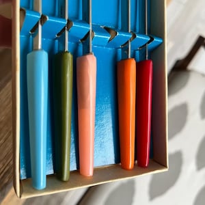 May include: A set of six vintage crochet hooks with colorful handles. The handles are in shades of blue, green, pink, orange, and red. The hooks are stored in a blue and tan cardboard box.