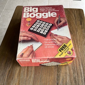 May include: A red and white box for the board game Big Boggle. The box features a close-up of the game board with letter tiles and a hand writing on a notepad. The text on the box reads "Parker Brothers Bigger Challenge 3-Minute Word Game Big Boggle".