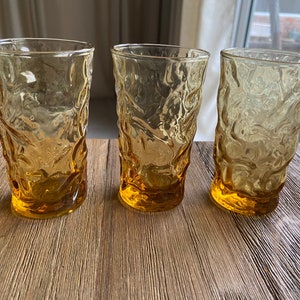 May include: Three amber-colored glass tumblers with a textured, rippled surface. The glasses are empty and sitting on a wooden surface.