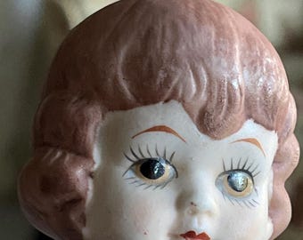 May include: A vintage doll head with a painted face and short, reddish-brown hair. The doll has detailed facial features, including painted eyebrows, eyelashes, and red lips. The doll's porcelain skin is pale, and the eyes are a dark brown color.