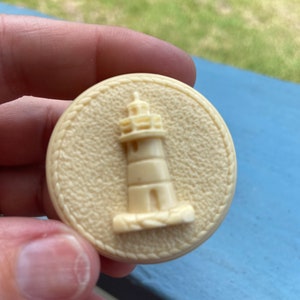 May include: A white ceramic button with a raised image of a lighthouse. The button has a rope-like border.