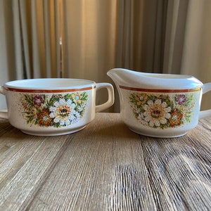 May include: A white ceramic creamer and sugar bowl set with a floral pattern. The creamer has a handle on the right side and the sugar bowl has two handles.