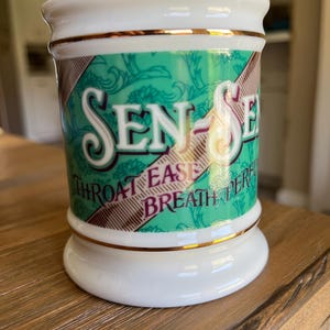 May include: A white ceramic mug with gold trim and the words "SEN-SE" in a decorative font. The mug has a green and brown patterned design with the words "THROAT EASE BREATH PERF" printed on it. The mug is sitting on a wooden surface.