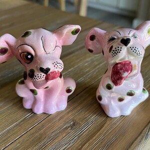 May include: Two pink ceramic dog figurines with black and green spots. The dogs have their tongues out and are painted with black noses and eyes. The figurines are on a wooden surface.