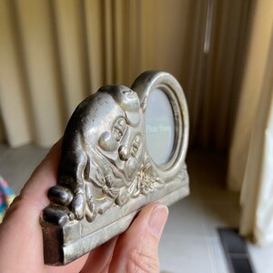 May include: Silver-toned photo frame with a cherub design. The frame has a small oval opening for a photo and the words "Photo Frame" are inscribed on the glass.