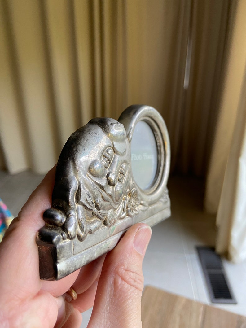 May include: Silver-toned photo frame with a cherub design. The frame has a small oval opening for a photo and the words "Photo Frame" are inscribed on the glass.