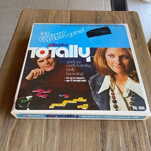 May include: A blue and white game box for the board game "Totally" by Aladdin's. The box features a photo of two people playing the game and the text "The great new numbers game!"