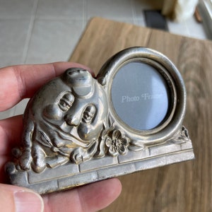 May include: Silver photo frame shaped like a cartoon face with a round opening for a photo. The frame has a brick base and floral accents. The text "Photo Frame" is printed on the glass.
