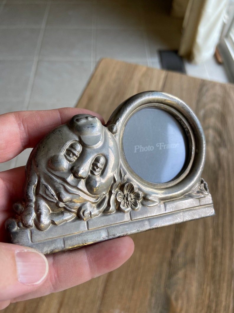 May include: Silver photo frame shaped like a cartoon face with a round opening for a photo. The frame has a brick base and floral accents. The text "Photo Frame" is printed on the glass.