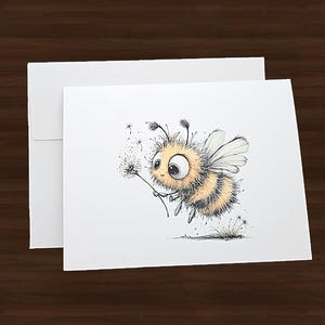 May include: A whimsical greeting card featuring a cartoon bee holding a dandelion. The bee is yellow and black with large eyes and wings. The card is white, with a matching envelope. The background is a dark brown wood.