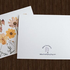 May include: Two white greeting cards. One card features pressed flowers in shades of yellow, orange, and purple. The other card is blank with the text "Willow Tree Papiery" and a website address. The cards are on a dark wood surface.