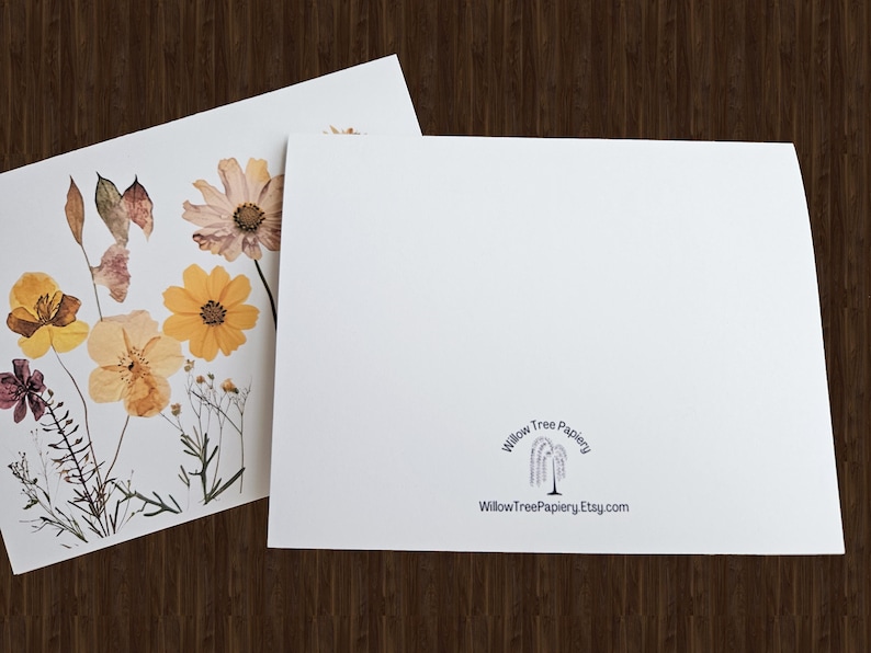 May include: Two white greeting cards. One card features pressed flowers in shades of yellow, orange, and purple. The other card is blank with the text "Willow Tree Papiery" and a website address. The cards are on a dark wood surface.
