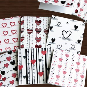 May include: A collection of greeting cards with various heart designs in red, black, and white. The cards feature patterns of hearts on strings, striped hearts, and simple heart outlines, perfect for Valentine's Day or expressing affection.