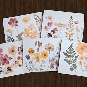 May include: Five square art prints featuring pressed flowers and foliage in shades of pink, yellow, and orange. The botanical arrangements are set against a white background, with a dark wood grain backdrop.