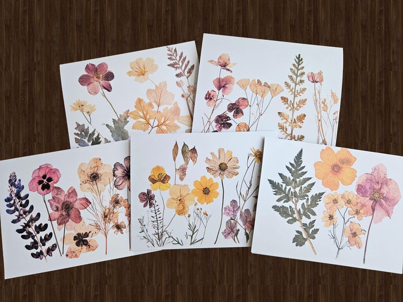 May include: Five square art prints featuring pressed flowers and foliage in shades of pink, yellow, and orange. The botanical arrangements are set against a white background, with a dark wood grain backdrop.