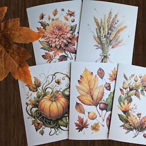 May include: A collection of five greeting cards featuring watercolor illustrations of autumn themes. The cards depict a large dahlia, a bundle of wheat, a pumpkin, leaves and acorns, and a wreath. The color palette includes warm tones of orange, brown, and gold.