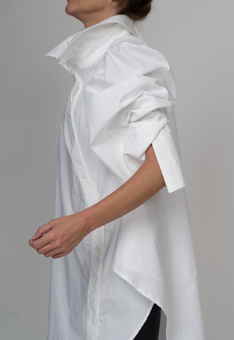 May include: A white oversized shirt with a high collar and long, voluminous sleeves. The shirt has a button-up front and a slightly asymmetrical hem.