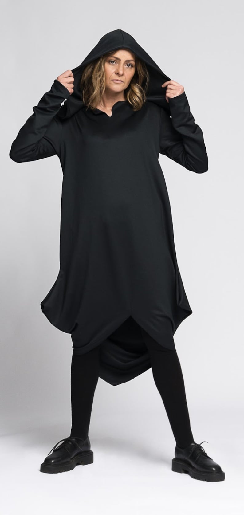 Op de afbeelding: Een zwarte kapjurk met een lange, vloeiende silhouet. De jurk heeft een gedrapeerd ontwerp en een ontspannen pasvorm. Het model draagt zwarte panty's en zwarte schoenen.