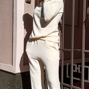 K&ouml;nnte beinhalten: Ein wei&szlig;er Strickpullover und eine passende Hose. Der Pullover hat einen Rollkragen und lange &Auml;rmel. Die Hose hat eine lockere Passform und ist an den Kn&ouml;cheln umgeschlagen. Die Person tr&auml;gt wei&szlig;e Turnschuhe.