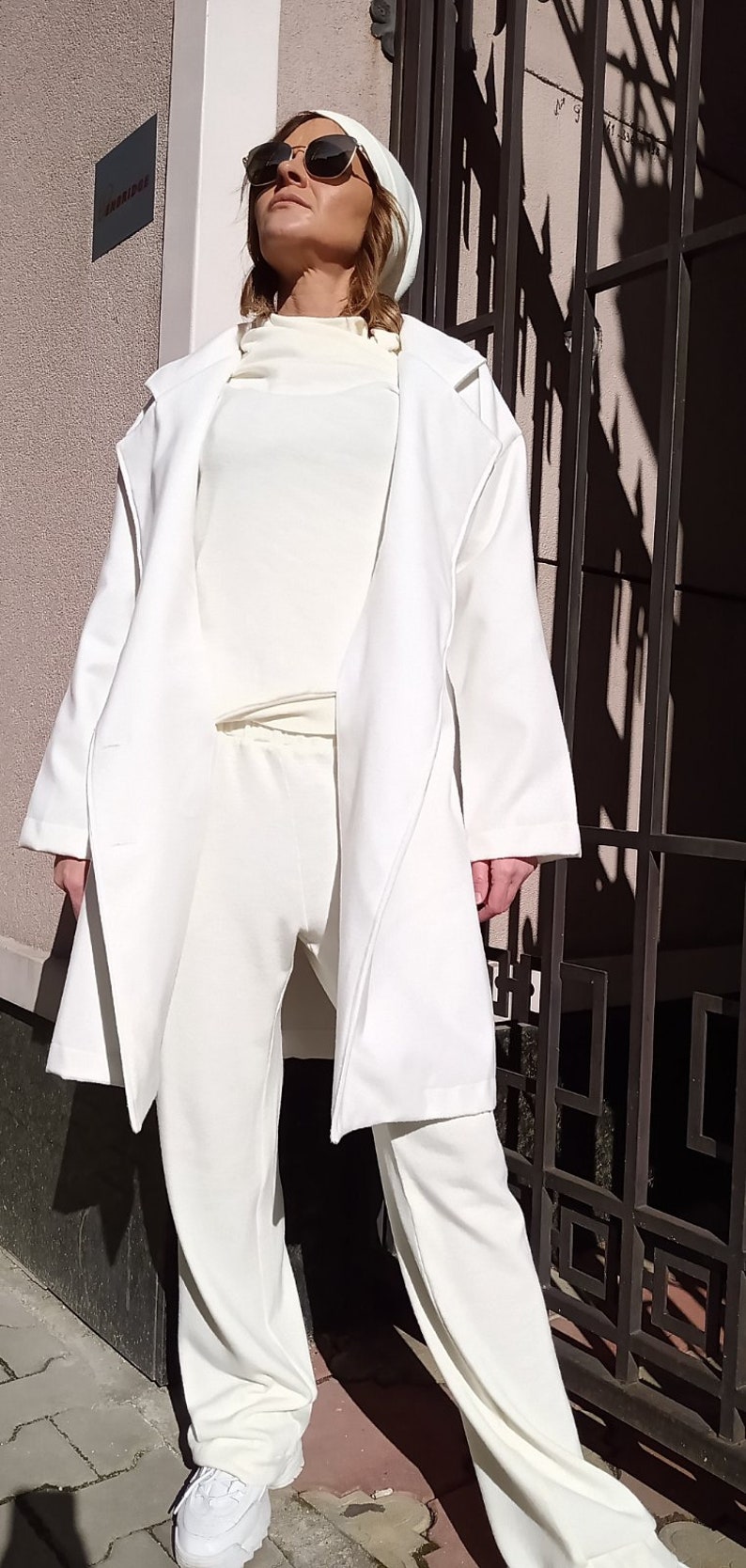 K&ouml;nnte beinhalten: Ein wei&szlig;er Langarmmantel mit einem wei&szlig;en Rollkragenpullover und einer wei&szlig;en Hose. Die Person tr&auml;gt eine Sonnenbrille und eine wei&szlig;e M&uuml;tze.