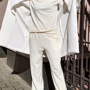 K&ouml;nnte beinhalten: Ein wei&szlig;es Outfit mit Hut, Hose und Pullover. Das Outfit wird von einer Person getragen, die vor einem wei&szlig;en Zaun steht.
