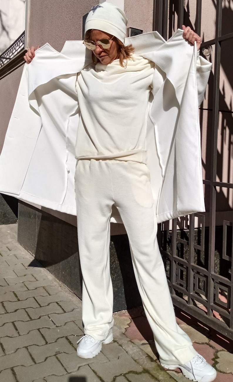 K&ouml;nnte beinhalten: Ein wei&szlig;es Outfit mit Hut, Hose und Pullover. Das Outfit wird von einer Person getragen, die vor einem wei&szlig;en Zaun steht.