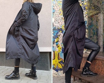 May include: A long black hooded raincoat with a zipper closure and pockets. The raincoat is worn by a person with black pants and black shoes.