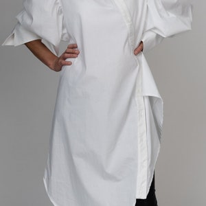 May include: White oversized shirt with a long, asymmetrical hemline and large, puffed sleeves. The shirt has a button-up front and a pointed collar.
