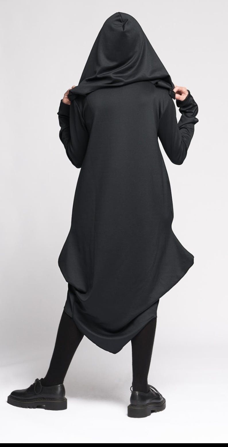 Op de afbeelding: Zwarte kap-tuniek met een lang, vloeiend ontwerp. De tuniek heeft een losse pasvorm en een geplooide zoom. Het model draagt zwarte panty's en zwarte schoenen.