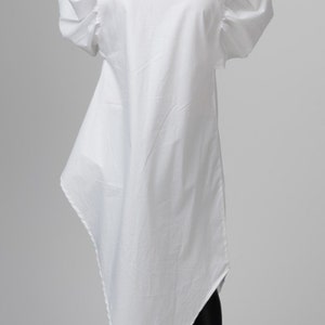 May include: A white long-sleeved shirt with a high collar and a triangular hemline. The shirt has puffed sleeves and is worn with black leggings and silver sneakers.