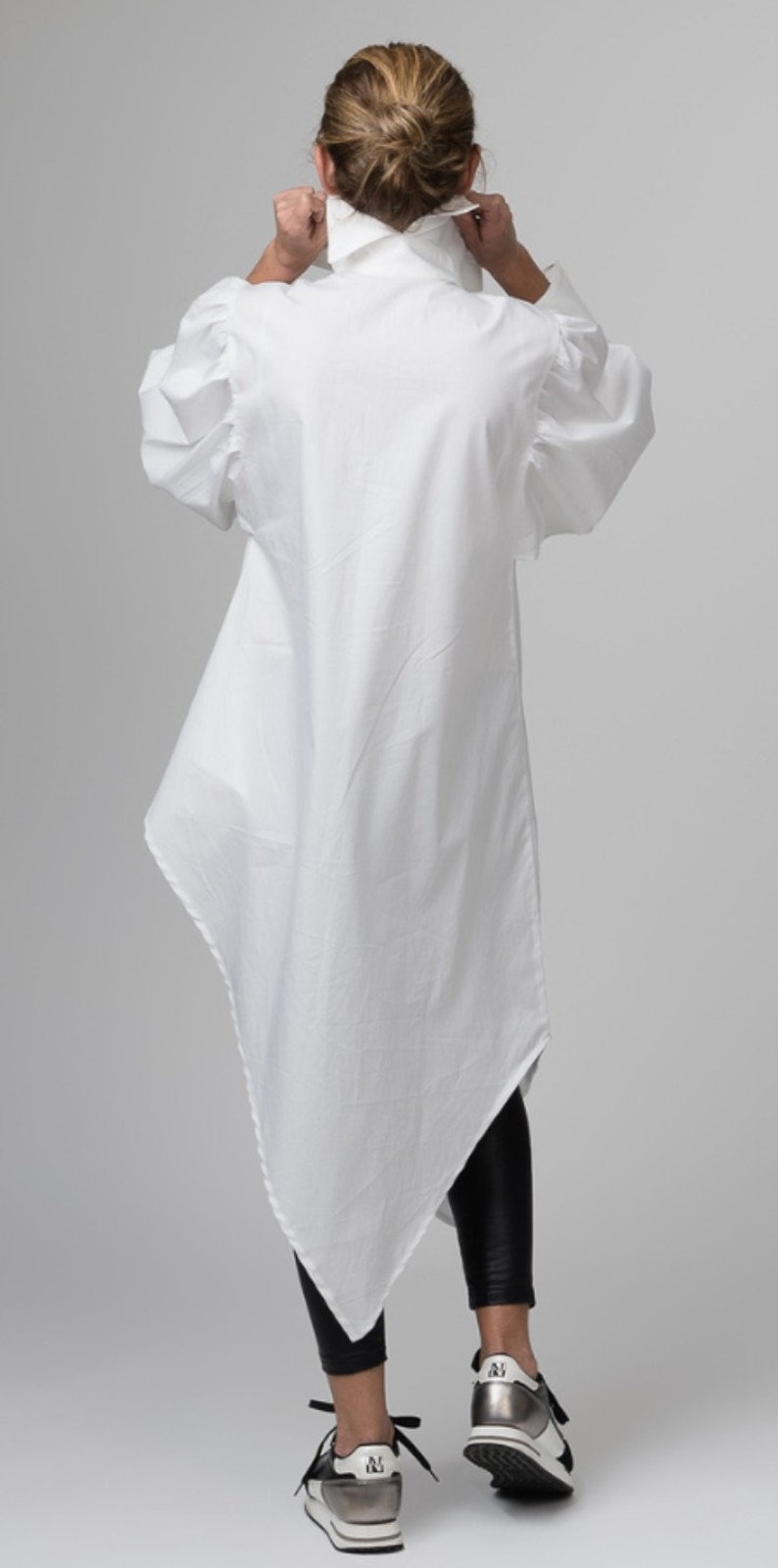 May include: A white long-sleeved shirt with a high collar and a triangular hemline. The shirt has puffed sleeves and is worn with black leggings and silver sneakers.