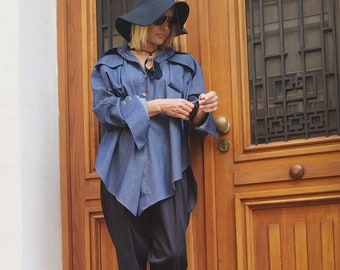 May include: A woman wearing a blue denim shirt with a black hat and black pants. The shirt has a unique asymmetrical design and the sleeves are rolled up. She is standing in front of a wooden door.