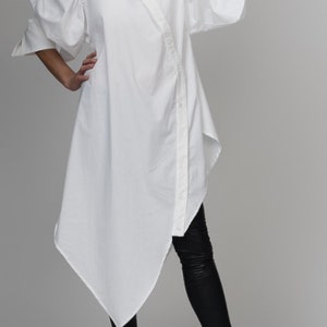 May include: A white, long-sleeved shirt with a button-up front and large, puffed sleeves. The shirt is asymmetrical, with a longer back hem and a shorter front hem. The model is wearing black leggings and black sneakers.