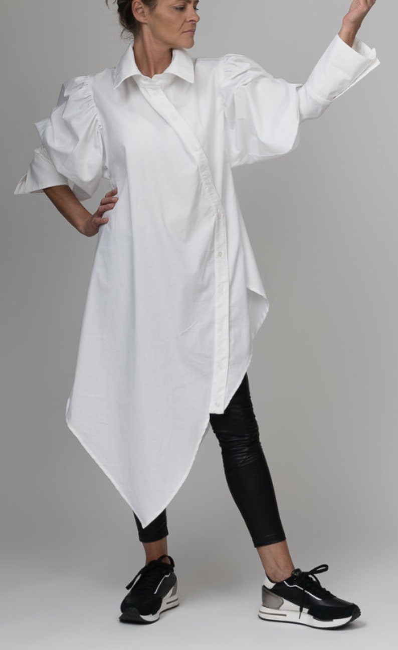 May include: A white, long-sleeved shirt with a button-up front and large, puffed sleeves. The shirt is asymmetrical, with a longer back hem and a shorter front hem. The model is wearing black leggings and black sneakers.
