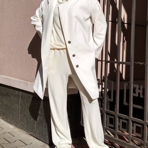 K&ouml;nnte beinhalten: Ein wei&szlig;er Langarmmantel mit Stehkragen und drei Kn&ouml;pfen. Der Mantel wird &uuml;ber einem wei&szlig;en Rollkragenpullover und einer wei&szlig;en Hose getragen. Die Person tr&auml;gt wei&szlig;e Turnschuhe.
