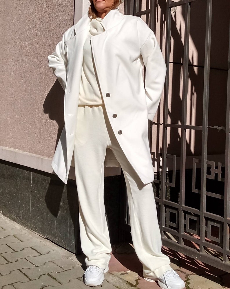 K&ouml;nnte beinhalten: Ein wei&szlig;er Langarmmantel mit Stehkragen und drei Kn&ouml;pfen. Der Mantel wird &uuml;ber einem wei&szlig;en Rollkragenpullover und einer wei&szlig;en Hose getragen. Die Person tr&auml;gt wei&szlig;e Turnschuhe.