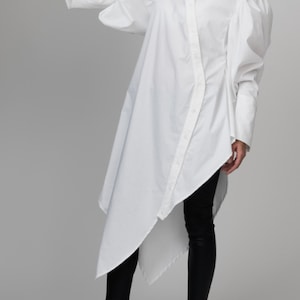 May include: A white oversized shirt with long sleeves and a button-up front. The shirt has a  asymmetrical hemline and puffed sleeves. The model is wearing black leggings and black sneakers.