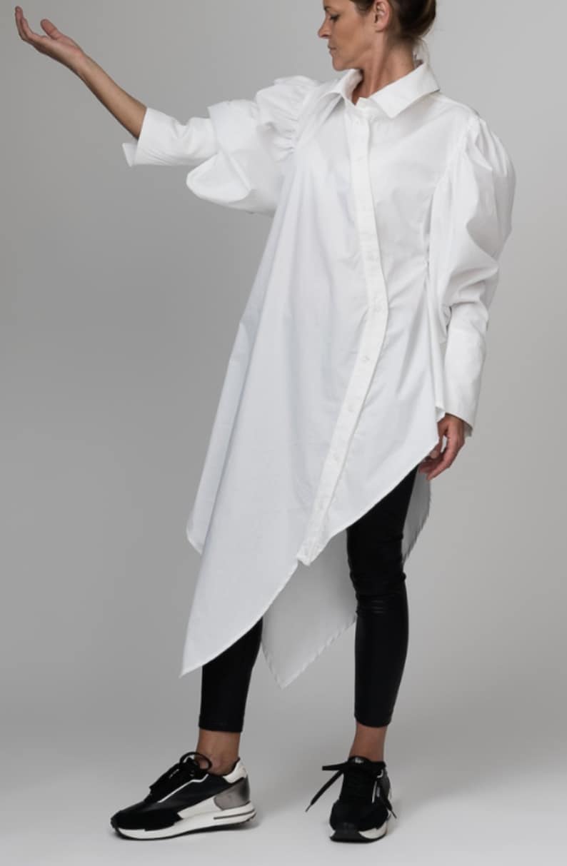 May include: A white oversized shirt with long sleeves and a button-up front. The shirt has a  asymmetrical hemline and puffed sleeves. The model is wearing black leggings and black sneakers.