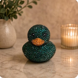 May include: A decorative duck figurine completely covered in small, sparkling dark green crystals. The duck features a distinct, rounded beak encrusted with bright orange crystals and two small, dark, circular eyes set within the green surface.