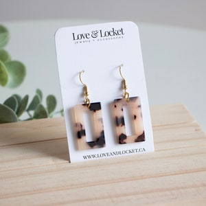 May include: A pair of gold tone earrings with rectangular tortoise shell-patterned resin dangles. The earrings have a simple hook closure. The earrings are displayed on a white card with the text "Love & Locket jewellery + accessories" and "www.loveandlocket.ca".