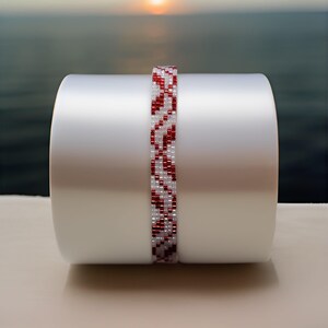 May include: A beaded bracelet with a repeating pattern of red and white beads. The bracelet is displayed on a white cylindrical object, with a blurred background of a sunset over the ocean.