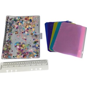 Op de afbeelding: Een transparante planner in ordnerstijl met kleurrijke confetti en een transparante sluiting. Daarnaast liggen verschillende gekleurde ordnerzakken in blauw, groen en roze. Een witte liniaal is ook inbegrepen.