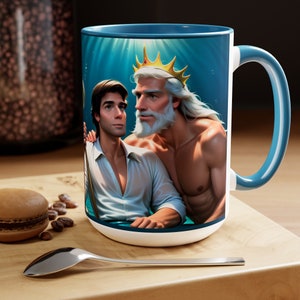 May include: A white ceramic mug with a blue rim and handle. The mug features a colorful illustration of a man with a white beard and a crown, and a man with brown hair, both underwater. The illustration is surrounded by a blue background with light blue rays.