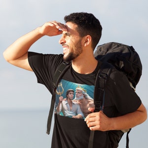 Puede incluir: Una camiseta negra con un estampado gráfico de dos hombres en el océano. El hombre de la izquierda sostiene un tridente y el hombre de la derecha lleva una corona. El texto en la camiseta dice "King Triton y su hijo".