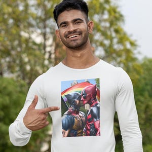 Puede incluir: Camiseta blanca de manga larga con un estampado gráfico de dos superhéroes besándose frente a una bandera arcoíris.