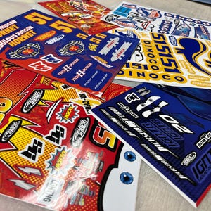 以下が含まれることがあります： レーシングカーのデザインが施された、カラフルなステッカーシートのコレクションです。「Fabulous Hudson Hornet」、「Twin H-Power」、「Rust-eze」などの車のグラフィック、数字、テキストが特徴です。主な色は赤、青、黄、白です。
