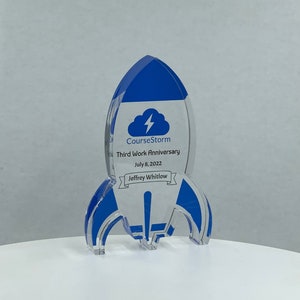 Puede incluir: Un premio de acrílico transparente azul y blanco en forma de cohete con el texto "CourseStorm Tercer aniversario de trabajo 8 de julio de 2022 Jeffrey Whitlow" en la parte delantera.