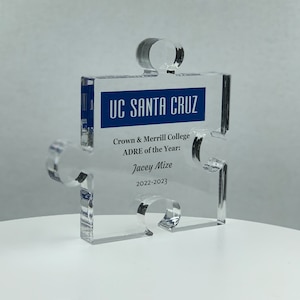May include: A clear plastic puzzle piece award with a blue banner that reads "UC SANTA CRUZ". The text below the banner reads "Crown & Merrill College ADRE of the Year: Jacey Mize 2022-2023".
